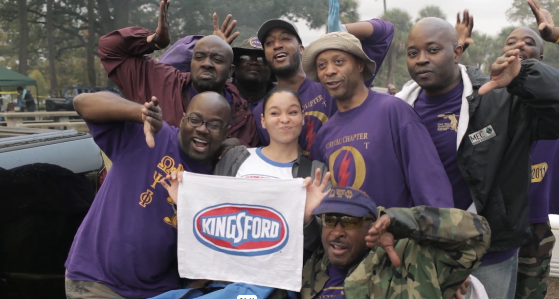 a group of people huddled together, holding up a Kingsford sign and smiling